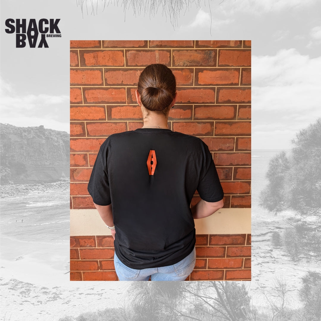 Person wearing a black t-shirt with an orange logo against a brick wall, with 'Shack Bay' branding.