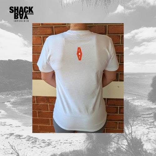 Person wearing a white shack bay brewing t-shirt with a orange logo on the back, standing against a brick wall.
